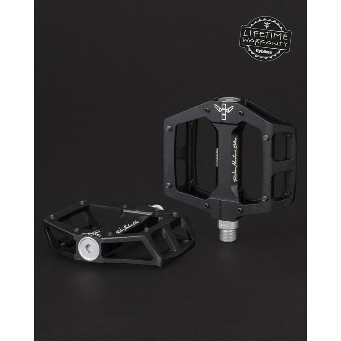 Two black Fly Bikes Ruben Aluminum Pedals with silver detailing are set against a dark backdrop. One is upright, highlighting its CNC Chromoly axle. The other is flat. A "Lifetime Warranty" logo in the top right promises durability with precision bearings.