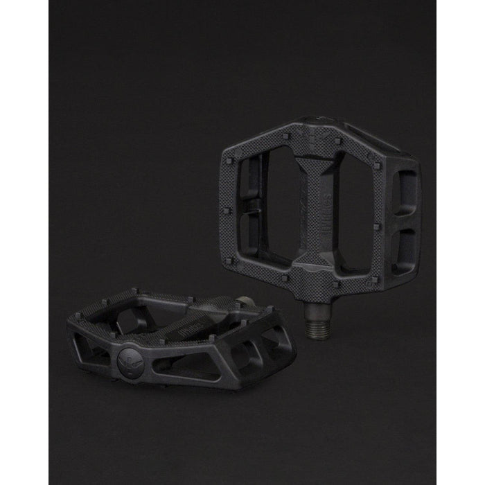 The Fly Bikes Ruben Pedals in Graphite are displayed on a dark backdrop, featuring two black pedals made from a nylon fiberglass compound. One is upright, and the other is on its side, showcasing textured surfaces and chromoly axle threads.