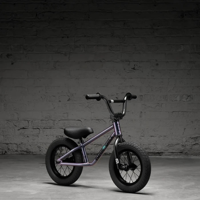 A compact purple Kink Coast 12 Inch Balance Bike (2026) with thick tires effortlessly leans against a textured gray brick wall on concrete, embodying its rugged style.