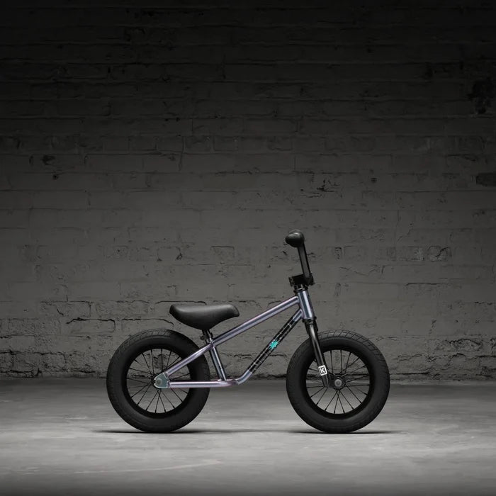 A Kink Coast 12 Inch Balance Bike (2026) with a purple frame and black seat, tires, and handlebars is positioned against a textured gray brick wall.