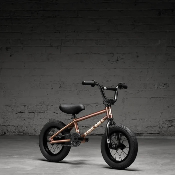 The Kink Roaster 12 Inch Bike (2026), ideal as a child's first BMX, boasts a bronze frame, black seat and wheels. It's set against a dark brick wall background and includes a reliable coaster brake for safe stopping.