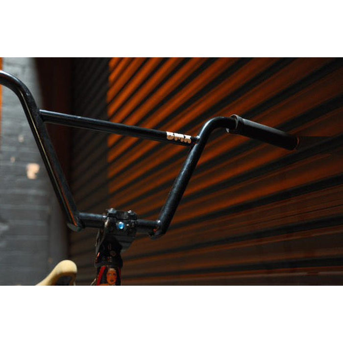 Close-up of Kink Williams 4-Piece Bars in black chromoly tubing, positioned before a slightly open brown garage door. Dim lighting casts shadows on the metal surface, emphasizing its sleek design.